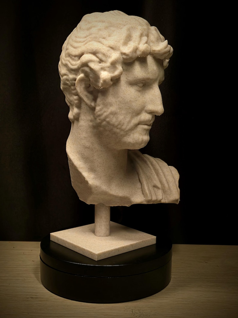 Puede incluir: Busto beige de un hombre con cabello rizado y barba, montado sobre una base cuadrada y un soporte cil&iacute;ndrico. La escultura se encuentra sobre un fondo negro, resaltando los detalles cl&aacute;sicos.
