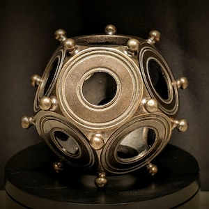 May include: A spherical bronze-toned object featuring a geometric pattern of recessed circular cutouts. The surface is adorned with raised spherical studs positioned at the intersections of the circular frames, creating a textured, multi-faceted appearance.