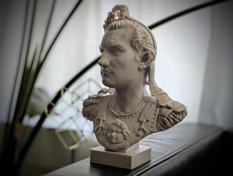 K&ouml;nnte beinhalten: Eine hellgraue B&uuml;ste einer r&ouml;mischen Figur, die einen detaillierten Brustpanzer mit einem zentralen Gesichtsemblem und einen Lorbeerkranz auf dem Kopf tr&auml;gt. Die Figur ist im Profil dargestellt und auf einem quadratischen Sockel montiert.