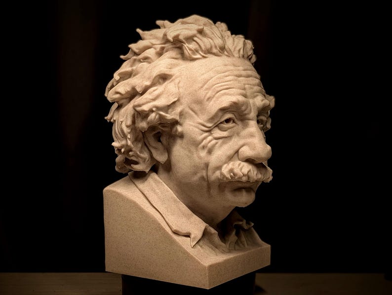 May include: A detailed bust of Albert Einstein, sculpted in a light beige material. The sculpture captures Einstein's iconic features, including his wild hair, mustache, and expressive face. The bust is set against a dark background.