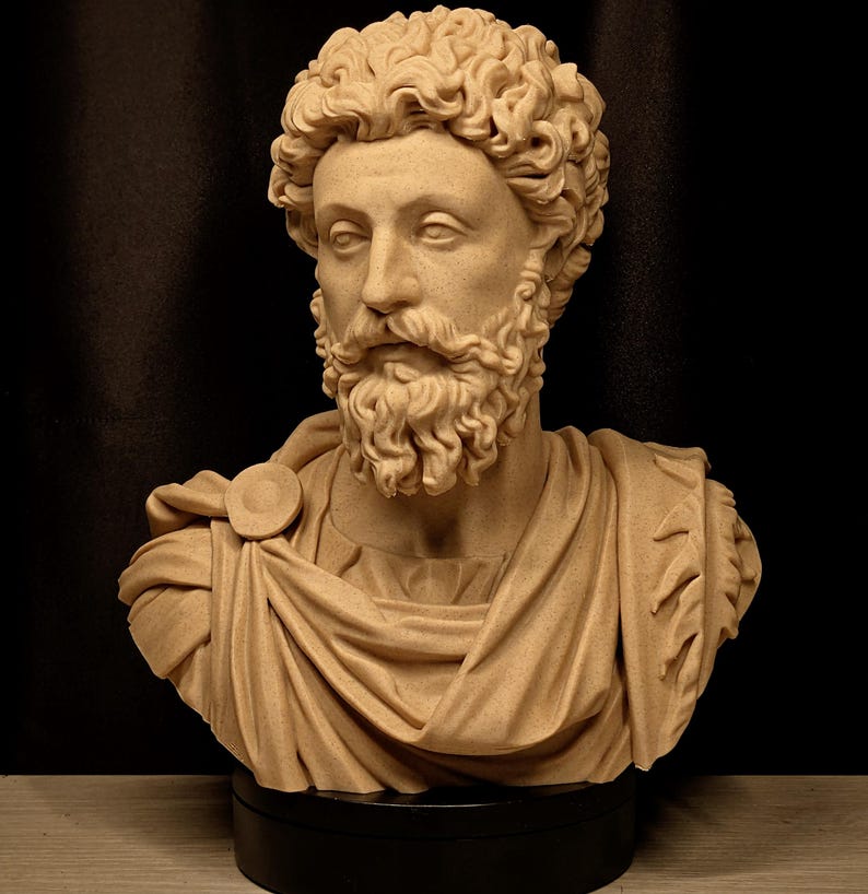 May include: A tan-colored bust of a man with curly hair and a beard. The sculpture depicts a Roman emperor, wearing a draped garment with a circular clasp. The bust is mounted on a black base, set against a dark background.