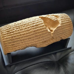 May include: A tan, cylindrical replica of the Cyrus Cylinder featuring detailed cuneiform inscriptions covering its surface, with a noticeable chipped section near the top, mounted on a simple black rectangular display stand.