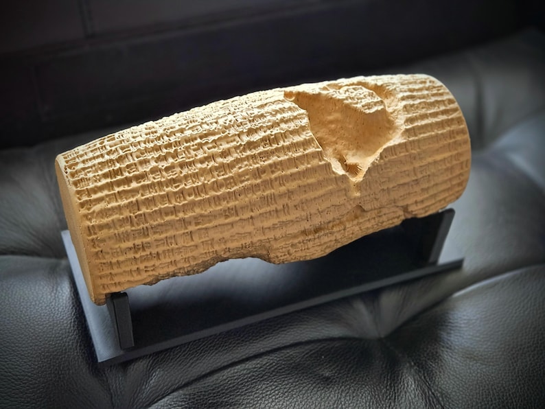 May include: A tan, cylindrical replica of the Cyrus Cylinder featuring detailed cuneiform inscriptions covering its surface, with a noticeable chipped section near the top, mounted on a simple black rectangular display stand.