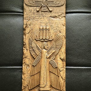 May include: A rectangular, brown relief carving featuring a winged figure with outstretched arms, wearing a long robe and a crown. Above the figure is a symbol with wings. The carving has detailed textures and inscriptions.