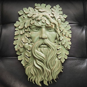 May include: A green-toned wall sculpture depicting a Green Man face, surrounded by oak leaves and acorns. The face has a long beard and detailed facial features. The sculpture is mounted on a black, textured surface.