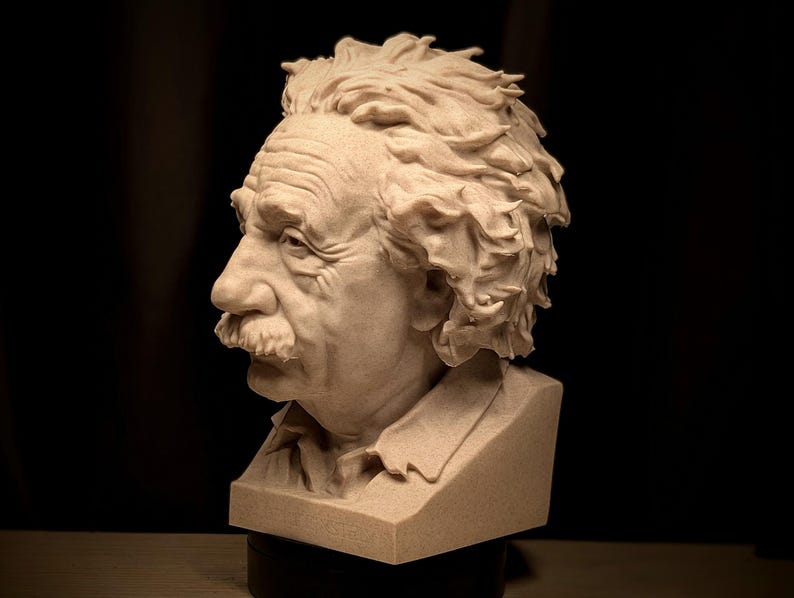 May include: A detailed bust sculpture of Albert Einstein, featuring his iconic hairstyle and mustache. The sculpture is in a light beige color, with visible details of his facial features and clothing. The bust is set against a dark background.