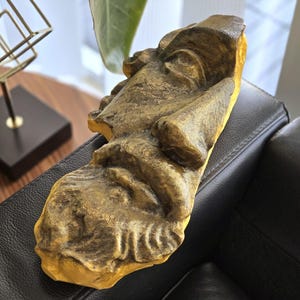 Puede incluir: Una escultura dorada de un rostro humano, posiblemente un fragmento de busto, descansa sobre una superficie de cuero negro. La escultura tiene detalles y una superficie texturizada, lo que sugiere una estética envejecida o antigua. El fondo incluye una obra de arte geométrica.