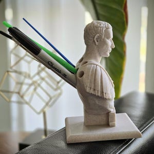 May include: A beige Roman emperor bust pen holder. It features a black Sharpie marker, a green pen, and a blue pen. The pen holder is on a square base. The bust is made of a stone-like material.