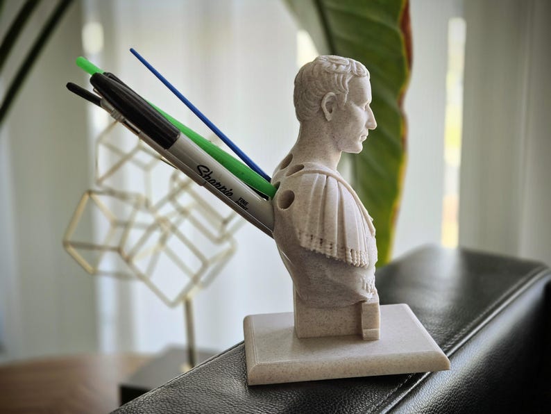 May include: A beige Roman emperor bust pen holder. It features a black Sharpie marker, a green pen, and a blue pen. The pen holder is on a square base. The bust is made of a stone-like material.