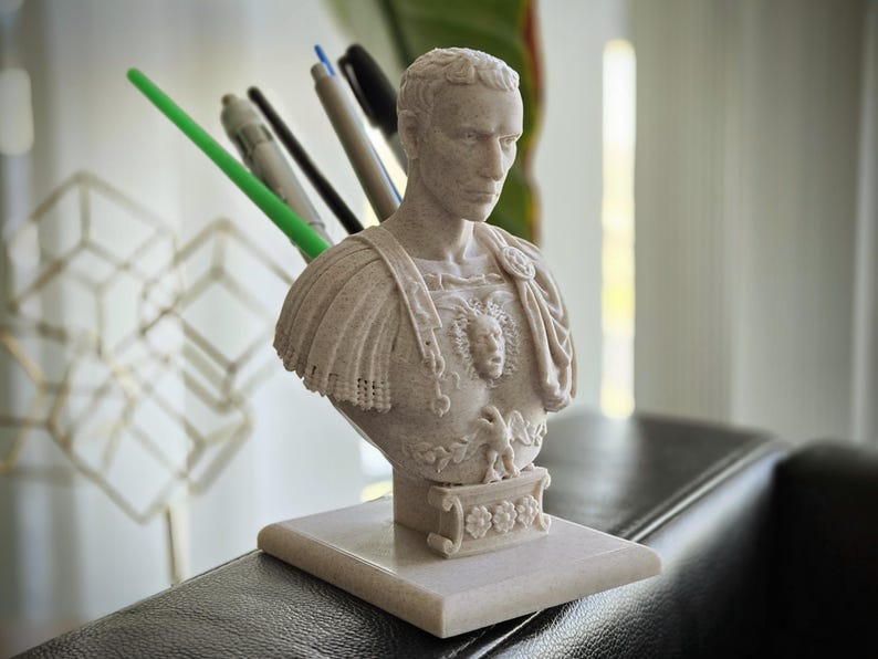 May include: A beige bust of a Roman emperor with a detailed design. The bust is mounted on a rectangular base and holds pens. The emperor's chest features a detailed emblem. The pens are green, black, and gray. The bust is on a dark surface.