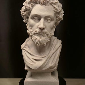 May include: A detailed bust sculpture of a man with curly hair and a beard. The sculpture is light beige and features a draped garment over the shoulders. It is mounted on a black base, set against a dark background.