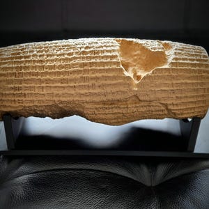 May include: A tan, cylindrical clay artifact featuring rows of cuneiform script etched into its surface. The object has a noticeable irregular chip on the upper right side and is displayed horizontally on a black, two-piece rectangular stand.