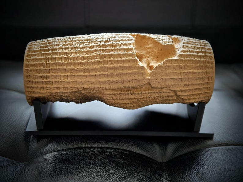 May include: A tan, cylindrical clay artifact featuring rows of cuneiform script etched into its surface. The object has a noticeable irregular chip on the upper right side and is displayed horizontally on a black, two-piece rectangular stand.