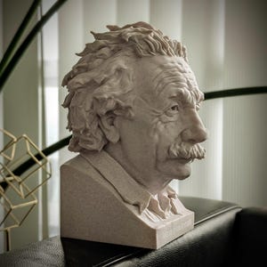 May include: A detailed bust of Albert Einstein, rendered in a light beige stone-like material. The sculpture captures Einstein's likeness with textured hair, a prominent mustache, and a thoughtful expression. The bust is set on a rectangular base.