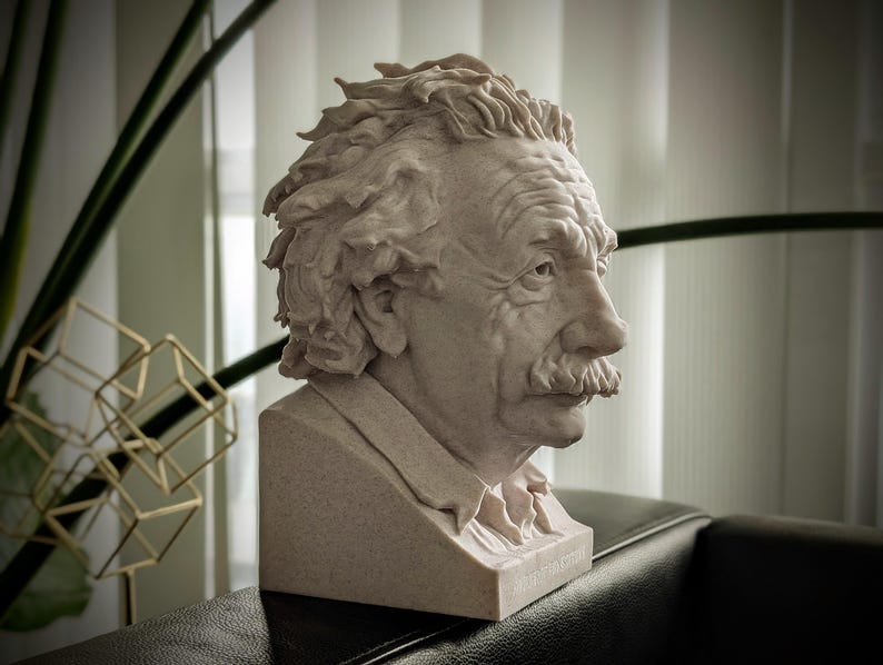 May include: A detailed bust of Albert Einstein, rendered in a light beige stone-like material. The sculpture captures Einstein's likeness with textured hair, a prominent mustache, and a thoughtful expression. The bust is set on a rectangular base.
