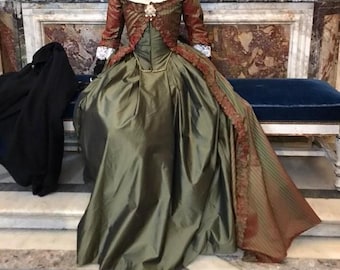 May include: A floor-length gown featuring a solid olive green skirt and a structured bodice with long sleeves in a rust-colored striped fabric. The bodice includes a square neckline, ruffled trim along the front opening, and white lace cuffs.