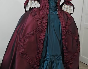 May include: A floor-length gown featuring a deep burgundy overcoat with ruffled trim and white lace cuffs, worn over a teal blue skirt with a ruffled hem. The bodice includes a teal blue panel with gathered fabric and a light pink choker necklace.