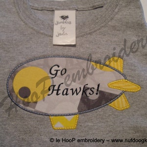 Puede incluir: Camiseta gris con un avión bordado en amarillo y gris que dice "Go Hawks!"