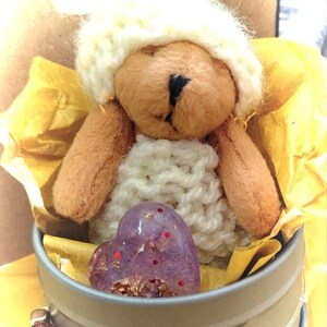 Op de afbeelding: Een handgemaakte teddybeer met een witte gebreide muts en trui, zittend in een kleine metalen container met een paarse hartvormige steen. Een kralenketting met roze en heldere stenen omringt de container.