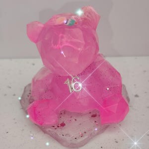 Könnte beinhalten: Eine pinkfarbene, facettierte Bärenfigur aus Kunstharz mit Glitzerakzenten. Der Bär sitzt auf einem passenden rosa-weißen Sockel. Ein silberner Anhänger mit der Zahl "16" ist am Hals des Bären befestigt. Die Figur ist ein Dekorationsartikel.