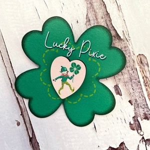 Pode incluir: Um trevo de quatro folhas verde com um coração rosa no centro. O coração apresenta uma ilustração de desenho animado de um duende segurando um trevo de quatro folhas. O trevo está etiquetado como "Lucky Pixie".