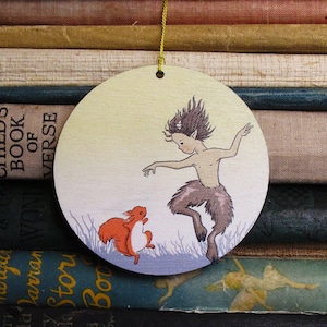 May include: A circular wooden decoration depicting a satyr dancing with a squirrel. The satyr has brown hair and fur, and the squirrel is orange and white. The ornament has a gold string for hanging.