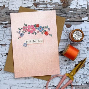 Può includere: Un biglietto di auguri rosa con illustrazioni floreali e la scritta "Just For You". Il biglietto è su una busta marrone, con accessori per cucire tra cui forbici, rocchetti di filo e un rocchetto di filo di legno.