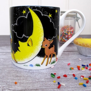 Puede incluir: Una taza de cerámica blanca con fondo negro y una luna creciente amarilla con una cara dormida. Un pequeño ciervo está de pie junto a la luna. La taza tiene un asa blanca.