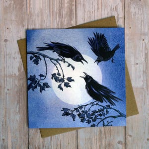 May include: A square greeting card featuring an illustration of three black crows against a large, bright full moon. Two crows are perched on dark, leafy branches, while one crow is in flight. The background is a textured, deep blue night sky.