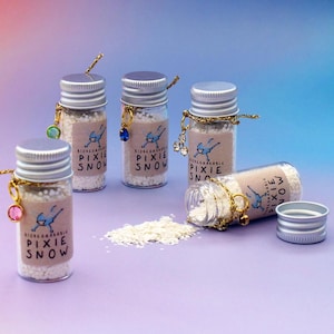 May include: Several small glass bottles filled with white granules, labelled "Biodegradable Pixie Snow." Each bottle has a silver lid and a charm. One bottle is on its side, with some of the contents spilled. The background is a gradient of purple and blue.