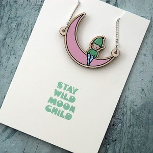 May include: A wooden necklace with a pink crescent moon pendant featuring a green elf sitting on the moon. The text "STAY WILD MOON CHILD" is printed on the card.