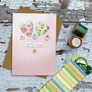 Könnte beinhalten: Eine handgefertigte Grußkarte mit zwei floralen Herzapplikationen auf rosa Hintergrund. Die Karte enthält den Text "With Love" und einen kleinen Knopf. Spulen mit grünem Faden und Stickgarn sind ebenfalls sichtbar.