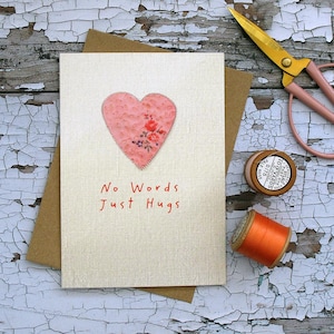 Könnte beinhalten: Eine handgefertigte Grußkarte mit einem rosa Herzen und den Worten "No Words Just Hugs" in Rot. Das Herz besteht aus floralem Stoff und ist auf die Karte genäht. Ein brauner Umschlag, eine Schere und Garnrollen sind ebenfalls zu sehen.