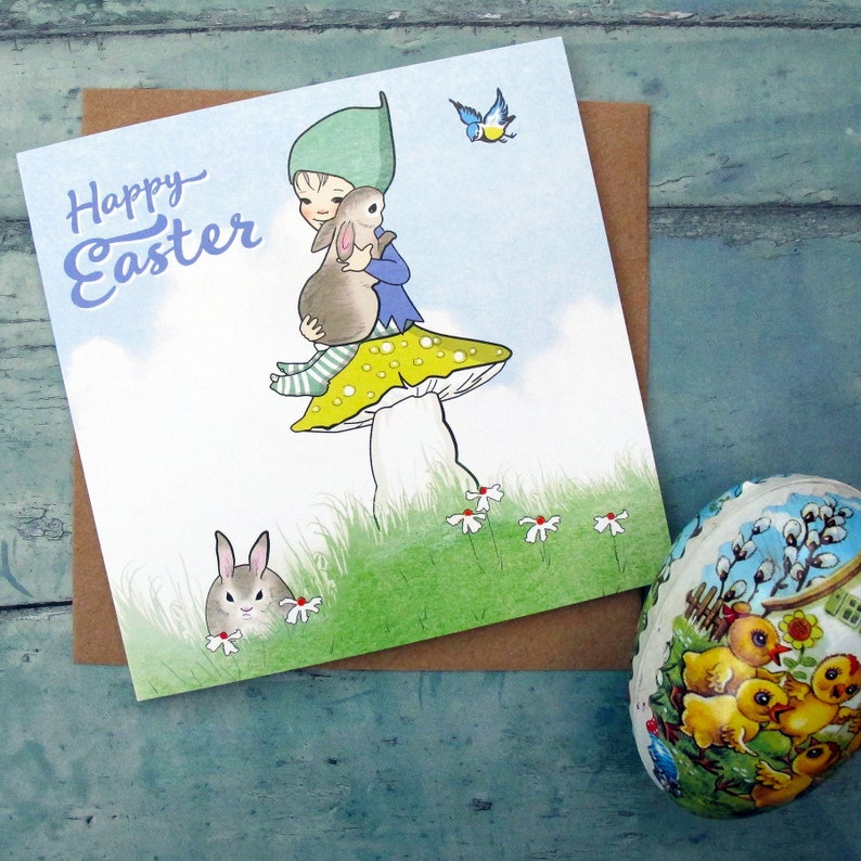 May include: A square greeting card featuring the text Happy Easter in blue script. The illustration shows a child in a green hat sitting on a yellow-spotted mushroom, holding a brown rabbit, with a second rabbit in the grass and a blue bird.