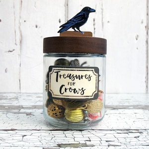 May include: A glass jar with a wooden lid and a decorative blue raven on top. The jar is labelled "Treasures for Crows" and filled with various trinkets. The jar sits on a white, distressed wooden surface.
