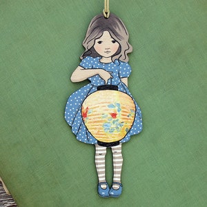 Könnte beinhalten: Ein Holz-Anhänger mit einer Mädchenfigur in einem blau-weiß gepunkteten Kleid, die eine gelbe und blaue Papierlaterne hält.