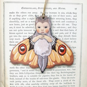 May include: A wooden ornament depicting a child dressed as a butterfly. The butterfly has orange and brown wings with a red eye on each wing. The child has a light brown complexion and is wearing a white dress with a black and white pattern. The ornament is hanging from a string.
