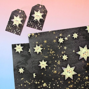 Puede incluir: Etiquetas de regalo y papel decorativo con temática celestial. Las etiquetas y el papel presentan un fondo oscuro con estrellas doradas y soles sonrientes. Los soles tienen un estilo de ilustración vintage con una cara y rayos.