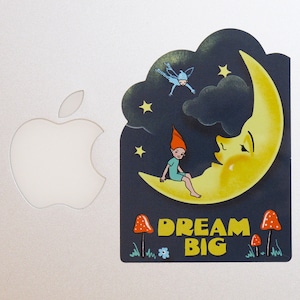 May include: A whimsical illustration of a crescent moon with a sleeping face, a small fairy flying above, and a gnome sitting on the moon. The moon is yellow with a dark blue background. The text "Dream Big" is written in white letters below the moon.