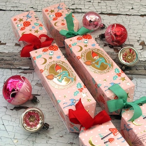 Puede incluir: Tres cajas de crackers de Navidad rosas y blancas con detalles dorados y un diseño de duendecillo caprichoso. Cada caja está atada con una cinta roja y verde.