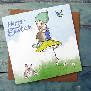 May include: A square greeting card featuring a cartoon illustration of a figure in a green hat sitting on a yellow-spotted mushroom while holding a brown rabbit. The text Happy Easter is printed in blue. A small blue bird and another rabbit are visible.