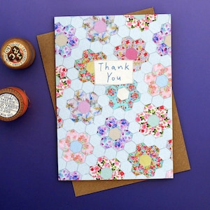 Puede incluir: Una tarjeta de felicitación hecha a mano con un estampado floral y las palabras "Thank You". La tarjeta presenta un fondo azul claro con formas hexagonales y diseños florales coloridos. La tarjeta está sobre un sobre marrón.
