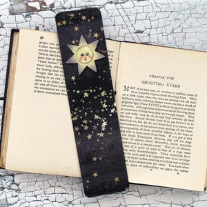 May include: A black bookmark with a celestial design featuring gold stars and a sun with a smiling face. The bookmark is placed on an open book with text visible.