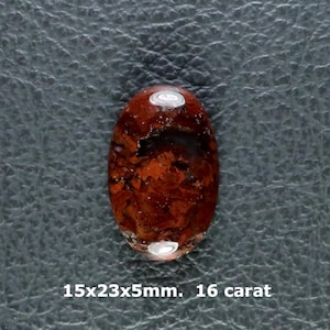 May include: An oval-shaped cabochon gemstone featuring a deep reddish-brown color with dark, mottled inclusions throughout. The stone measures 15x23x5mm and weighs 16 carat, with two small, circular, reflective silver-toned metallic accents.