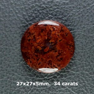 Pode incluir: Uma pedra preciosa polida de formato redondo, com uma cor castanho-avermelhada profunda e inclusões escuras complexas semelhantes a musgo. A pedra mede 27x27x5mm e pesa 34 quilates, apresentando um acabamento liso e reflexivo.