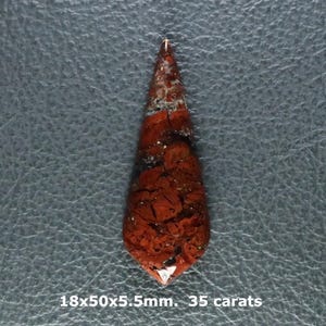May include: A teardrop-shaped red and black jasper gemstone cabochon with a polished surface. The stone features intricate dark veining and natural patterns. Text printed below the stone reads 18x50x5.5mm. 35 carats.