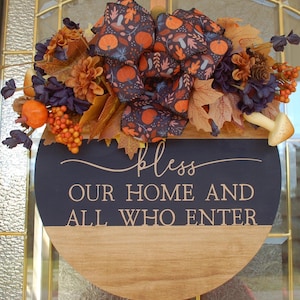 May include: A round wooden sign with a navy blue top and a natural wood bottom. The sign is decorated with fall foliage, a bow with pumpkins, and the text "bless OUR HOME AND ALL WHO ENTER".