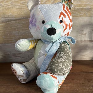 Peut inclure: Un ours en peluche patchwork fait à la main, composé de divers tissus aux motifs et couleurs variés. L'ours a un nez noir, des yeux en boutons et un ruban bleu clair autour du cou. Les tissus présentent des motifs floraux, géométriques et abstraits.