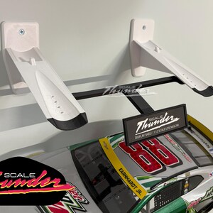 May include: White and black display shelf for a 1:12 scale NASCAR model car. The shelf is mounted on a wall and features the "Scale Thunder" logo. The model car has the number 88 and the name "Earnhardt Jr."