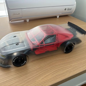 May include: A clear polycarbonate remote control car body shell featuring a red interior roll cage structure, black wheels with white Hoosier lettering, and a black rear spoiler. The car body is transparent and molded to fit a racing vehicle chassis.